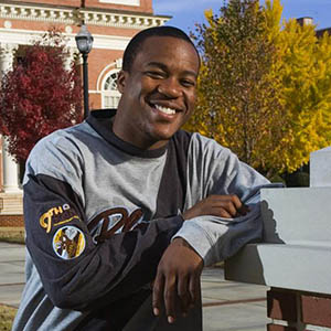 A smiling student leaning on a building column with his elbow looking at the camera