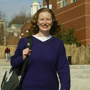 A student in a purple sweater wearing a book bag smiling at the camera as she walks towards it